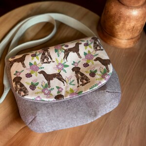 May include: A small, gray and white crossbody bag with a floral print featuring brown dogs. The bag has a light green strap.