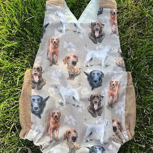 Pode incluir: Um macacão de bebê bege com acabamento marrom e um padrão de várias raças de cães. Os cães são retratados de forma realista e são cercados por pegadas.