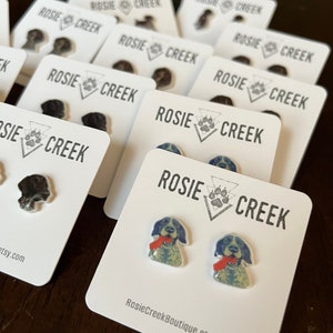 Pode incluir: Um conjunto de dois pares de brincos de botão com tema de cães. Um par apresenta um cão preto e branco com a língua vermelha para fora. O outro par apresenta um cão preto e branco com a língua preta para fora. Os brincos são embalados em cartões brancos com o texto "Rosie Creek" impresso neles.