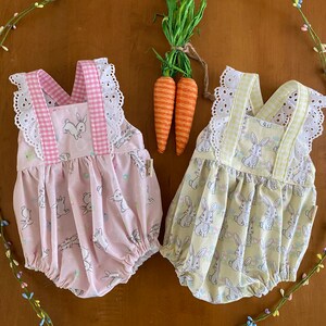 Könnte beinhalten: Zwei Baby-Strampler mit Hasenaufdrucken. Einer ist rosa mit Vichy-Besatz, der andere gelb mit Vichy-Besatz. Beide haben weiße Lochspitzenbesätze und Hasenmuster. Der Hintergrund umfasst dekorative Karotten und florale Akzente.