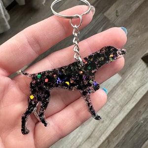 May include: Black and silver glitter dog keychain. The dog is a German Shorthaired Pointer breed. The keychain is made of resin and has a silver metal ring.