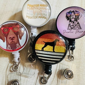 May include: Four different retractable badge holders with colorful designs featuring a dog breed. The designs include a white dog with the text "Pointer Mama" repeated in a gradient, a black and white dog wearing sunglasses, a black silhouette of a dog on a sunset background, and a brown and white dog wearing glasses.
