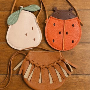 May include: Three small crossbody bags with thin straps. One is a light beige pear shape with a green leaf, one is a round orange ladybug shape with a brown top, and one is a round tan bag featuring a braided top edge and hanging fringe.