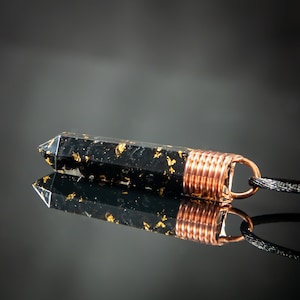 Könnte beinhalten: Ein schwarzer und goldener Anhänger mit einer Kupferdrahtumwicklung und einer schwarzen Schnur. Der Anhänger hat die Form eines langen, dünnen Rechtecks mit einer spitzen End.