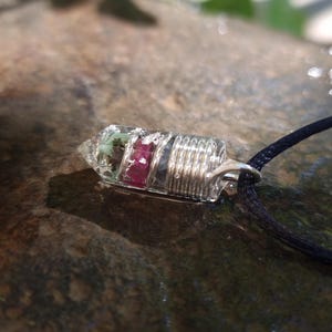 Könnte beinhalten: Ein mit Silberdraht umwickelter Kristallanhänger mit grünen und rosa Steinen im Inneren, der an einer schwarzen Schnur hängt.