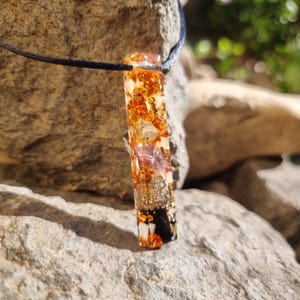 May include: A rectangular pendant necklace with a black cord. The pendant is a clear resin with embedded gold flakes, orange, purple, and black elements. The pendant is approximately 2 inches long.