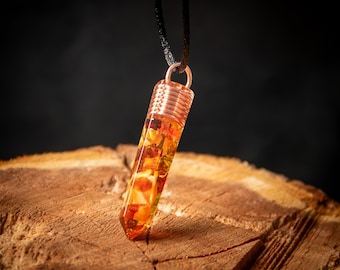 Puede incluir: Un colgante envuelto en alambre de cobre con resina transparente llena de flores y hojas secas. El colgante cuelga de un cordón negro y descansa sobre un trozo de madera.