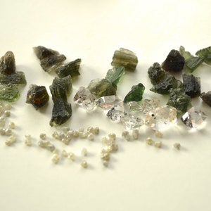 May include: A collection of green and brown rough gemstones and clear faceted crystals. The gemstones are scattered on a white surface.