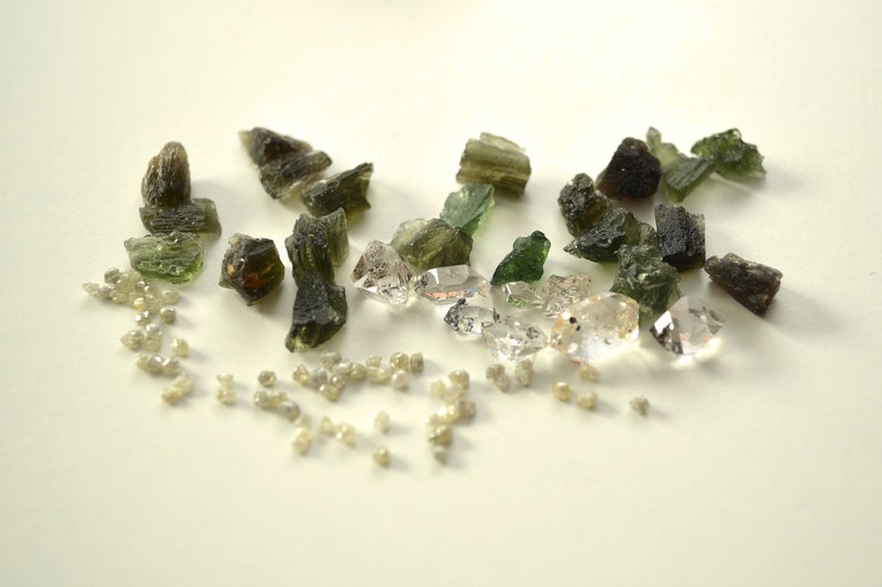 May include: A collection of green and brown rough gemstones and clear faceted crystals. The gemstones are scattered on a white surface.