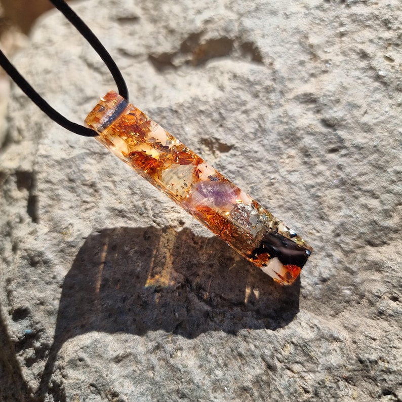 May include: A rectangular pendant necklace with a black cord. The pendant is a clear resin filled with various stones and metallic flakes in shades of orange, gold, and purple. The pendant is resting on a gray stone surface.