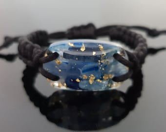 May include: A black braided bracelet with a blue and gold resin charm. The charm is oval shaped and has gold flecks throughout.