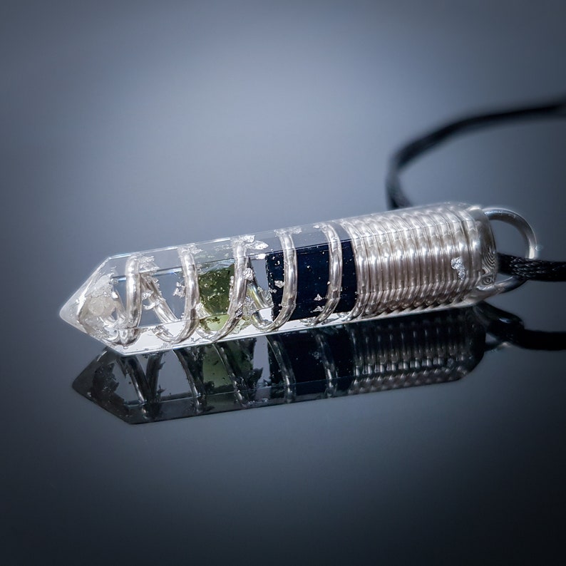 May include: A clear quartz crystal pendant with a silver wire coil and green and black stones inside. The pendant is attached to a black cord.