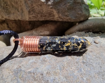 Puede incluir: Un collar con colgante negro y dorado con un envoltorio de alambre de color cobre. El colgante es un cristal con motas negras y doradas. El cordón del collar es negro.