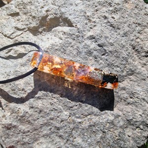 May include: A rectangular pendant with a black cord. The pendant is made of clear resin with embedded gold flakes, purple and clear stones, and a black and white design at the end. The pendant is resting on a gray rock.
