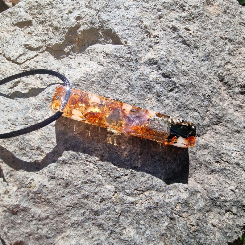 May include: A rectangular pendant with a black cord. The pendant is made of clear resin with embedded gold flakes, purple and clear stones, and a black and white design at the end. The pendant is resting on a gray rock.