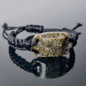 Könnte beinhalten: Ein schwarzes geflochtenes Armband mit einem klaren Harz-Anhänger, der goldene Flocken enthält.