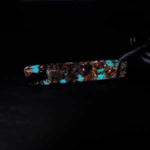 May include: A rectangular pendant with a dark background. The pendant is a mix of black, copper, and turquoise. The turquoise sections appear to glow. The pendant is attached to a dark cord.