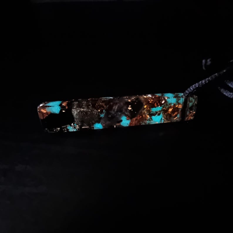 May include: A rectangular pendant with a dark background. The pendant is a mix of black, copper, and turquoise. The turquoise sections appear to glow. The pendant is attached to a dark cord.