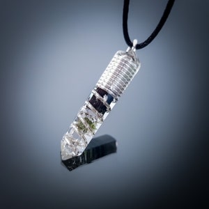 May include: A silver wire wrapped around a clear quartz crystal pendant with green and black inclusions. The pendant is attached to a black cord.