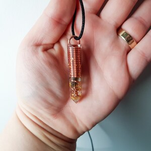 May include: A clear quartz crystal pendant with a copper wire coil inside. The pendant is on a black cord and has gold flecks throughout the crystal.