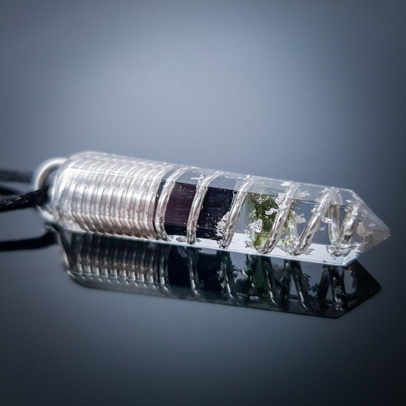 May include: A clear quartz crystal pendant with a silver wire wrapped around it. The crystal contains green and black inclusions. The pendant is attached to a black cord.