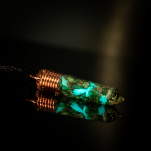 May include: A copper-wrapped pendant with a green and blue resin filled with small stones. The pendant has a pointed end and is reflecting light.