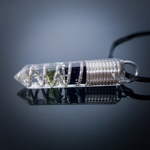 May include: A clear crystal pendant with a silver wire coil wrapped around a black stone and a green stone inside. The pendant is on a black cord.