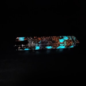 May include: A rectangular, clear resin object with copper flakes and glowing turquoise accents. The object is set against a black background, highlighting the internal details and the vibrant, luminous colors.