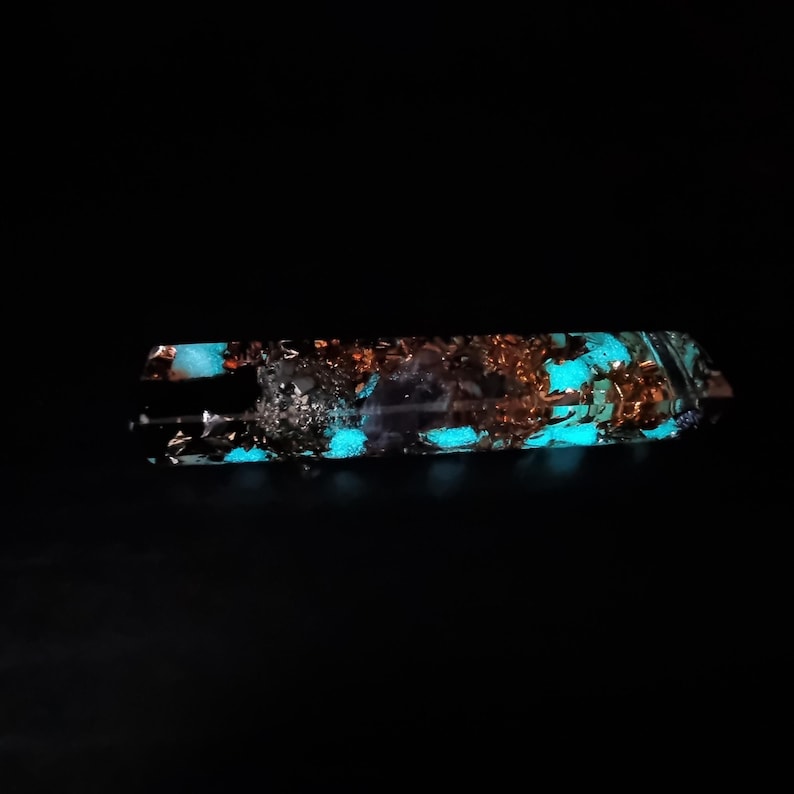 May include: A rectangular, clear resin object with copper flakes and glowing turquoise accents. The object is set against a black background, highlighting the internal details and the vibrant, luminous colors.