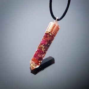 Op de afbeelding: Een hanger omwikkeld met koperdraad met een heldere hars gevuld met kleine rode en gouden stukjes. De hanger hangt aan een zwart koord.