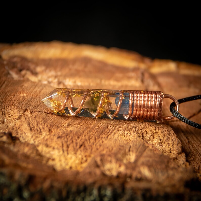 May include: A clear quartz crystal pendant with copper wire wrapped around it. The crystal is filled with gold flakes and a blue stone. The pendant is attached to a black cord.