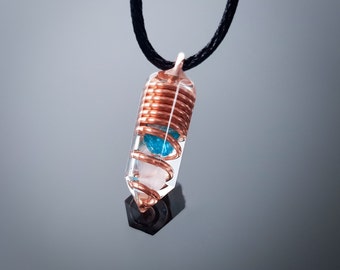 Op de afbeelding: Een heldere kristallen hanger met een koperen draad eromheen gewikkeld en een blauwe steen erin. De hanger is bevestigd aan een zwart koord.
