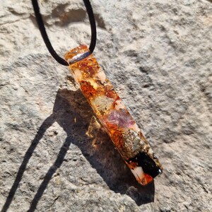 May include: A rectangular pendant necklace with a black cord. The pendant is made of clear resin with embedded gold flakes, orange and purple stones, and black accents. The pendant is approximately 2 inches long.