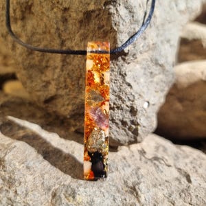 May include: A rectangular pendant necklace with a black cord. The pendant is a clear resin with embedded gold flakes, orange, purple, and black elements. The necklace is displayed on a gray rock surface.