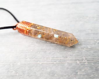 Op de afbeelding: Een hanger omwikkeld met koperdraad met een heldere hars punt gevuld met gouden vlokken en iriserende glitter. De hanger is bevestigd aan een zwart koord.