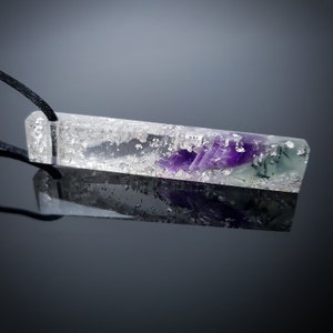 Könnte beinhalten: Ein klarer rechteckiger Anhänger mit einem violetten und grünen Kristall, der in Harz gefasst ist. Der Anhänger ist an einer schwarzen Schnur befestigt.
