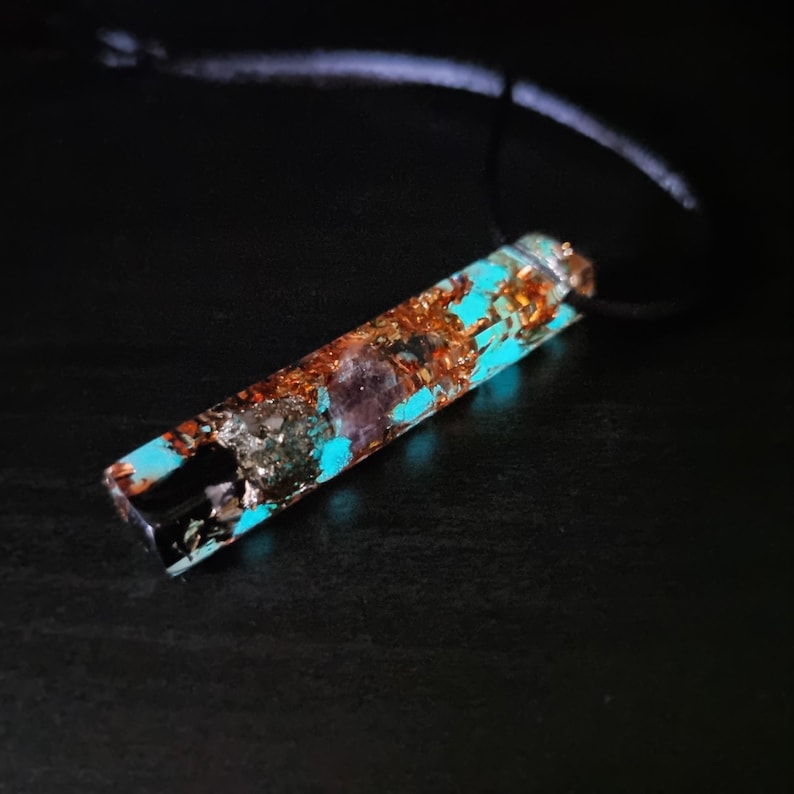May include: A rectangular pendant necklace with a black cord. The pendant is a mix of turquoise, copper, and purple hues, encased in clear resin. The pendant's design is a blend of natural elements and artistic craftsmanship.