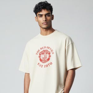 May include: Cream-colored t-shirt featuring a red crest with the text "THE RED DEVILS MANCHESTER UNITED EST 1970." The shirt has short sleeves and a relaxed fit. The graphic is centered on the chest.