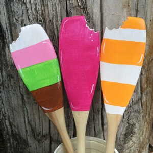 Puede incluir: Tres cucharas de madera pintadas para parecer paletas de helado. Las cucharas est&aacute;n pintadas de rosa, naranja y verde y rayas blancas. Las cucharas est&aacute;n dispuestas en una fila con la cuchara rosa en el medio.