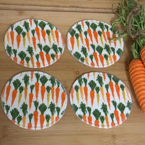 May include: Four round coasters with a white background and a repeating pattern of orange and yellow carrots with green tops. The coasters have a white stitched edge and are arranged on a wooden surface.