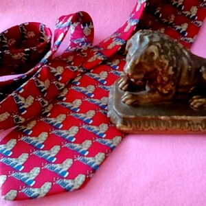 May include: A red silk tie with a repeating pattern of gold lions. The tie is folded and tied in a knot. A small bronze lion statue is sitting on a wooden base.