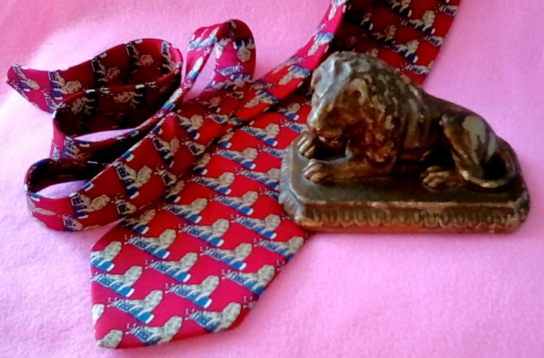 May include: A red silk tie with a repeating pattern of gold lions. The tie is folded and tied in a knot. A small bronze lion statue is sitting on a wooden base.