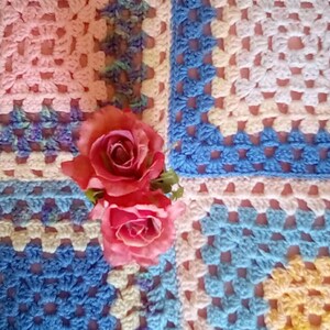 May include: A crocheted blanket made of squares in pink, blue, and yellow. The blanket is decorated with two pink roses.