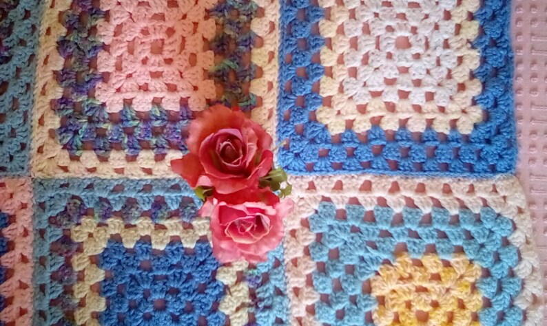 May include: A crocheted blanket made of squares in pink, blue, and yellow. The blanket is decorated with two pink roses.