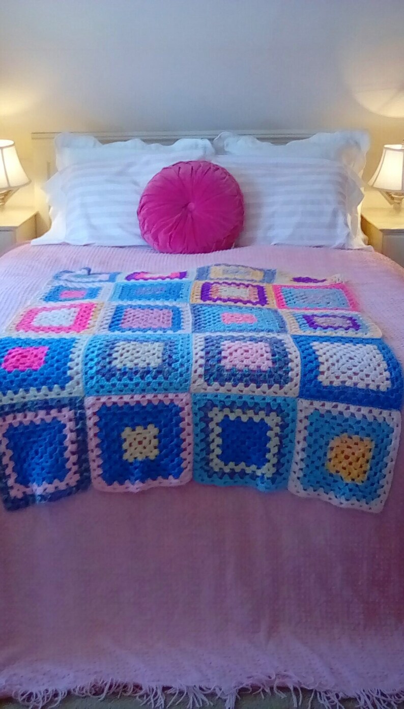 May include: A pink bed with a white and pink striped bedspread and a pink crocheted throw blanket with colorful squares. A pink round pillow is on top of the bedspread.