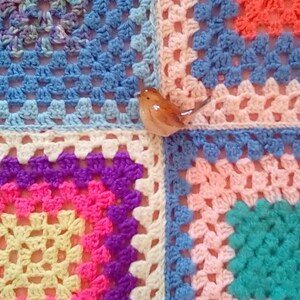 May include: A colorful crocheted blanket with a variety of squares in different colors, including blue, pink, yellow, purple, and green. A small ceramic bird figurine is placed on top of the blanket.