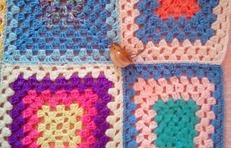 May include: A colorful crocheted blanket with a variety of squares in different colors, including blue, pink, yellow, purple, and green. A small ceramic bird figurine is placed on top of the blanket.