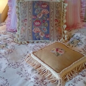 May include: A white bed with a pink and white striped pillow, a floral tapestry pillow, and a beige pillow with fringe. The bedspread has a floral pattern.