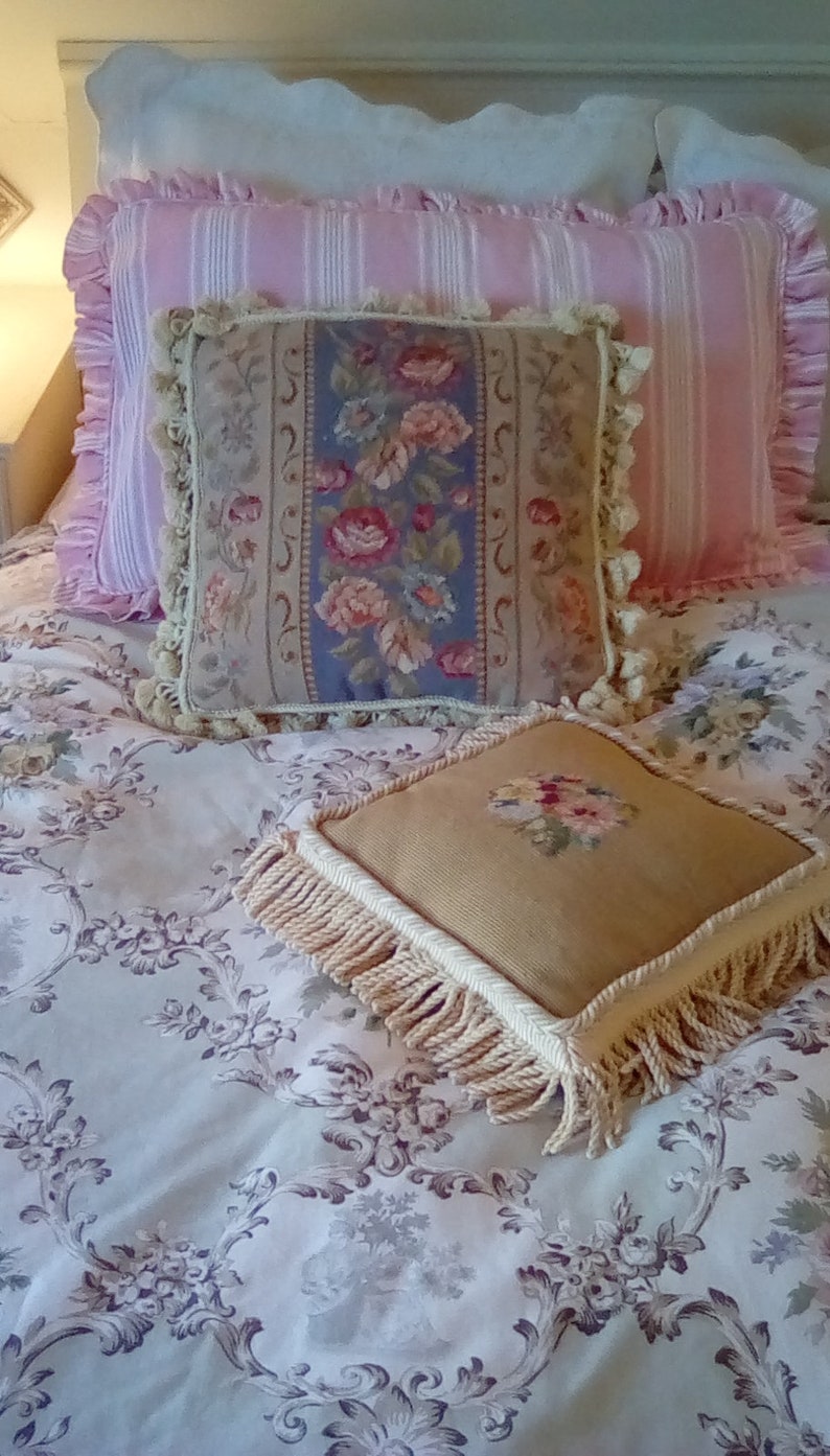 May include: A white bed with a pink and white striped pillow, a floral tapestry pillow, and a beige pillow with fringe. The bedspread has a floral pattern.