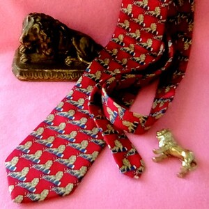 May include: A red silk tie with a repeating pattern of gold lions. The tie is lying on a pink surface with a gold lion statue.